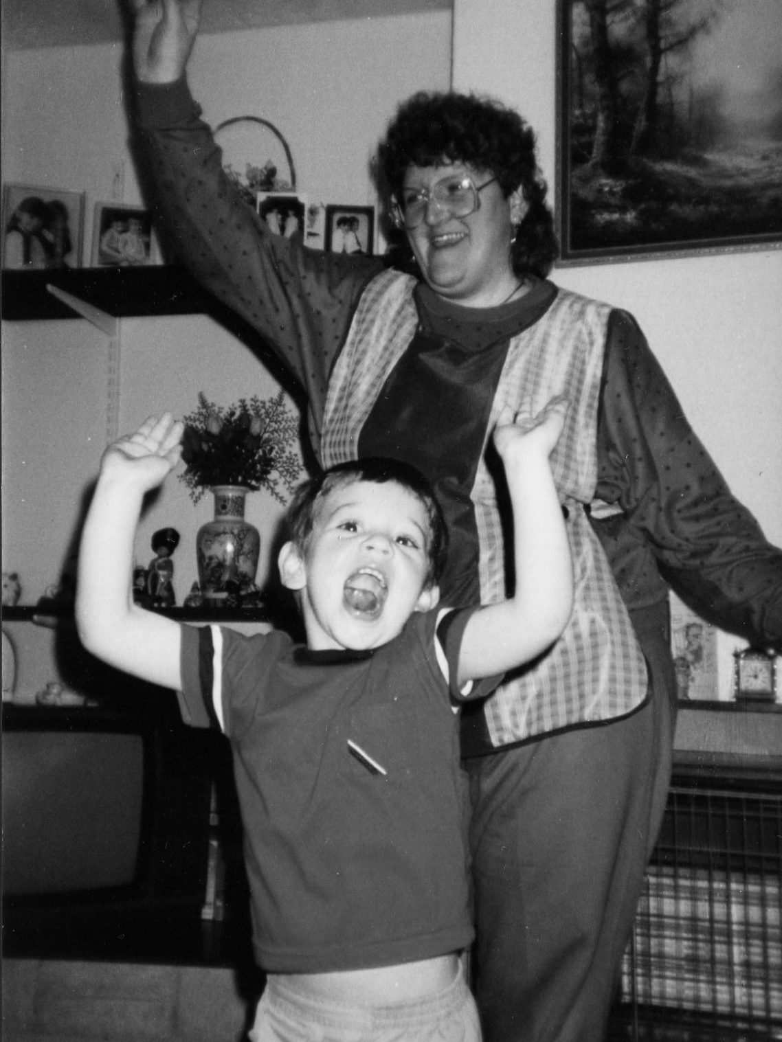 A black and white photo of an adult and child in front of a fireplace, both with one arm raised. Shelves behind them hold various objects.